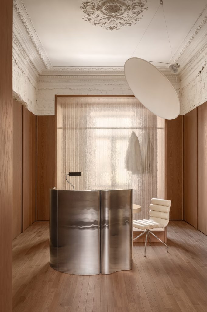 Wavy stainless-steel reception desk on oak flooring, oak-panelled walls and a restored ceiling frieze above, a disc pendant alongside