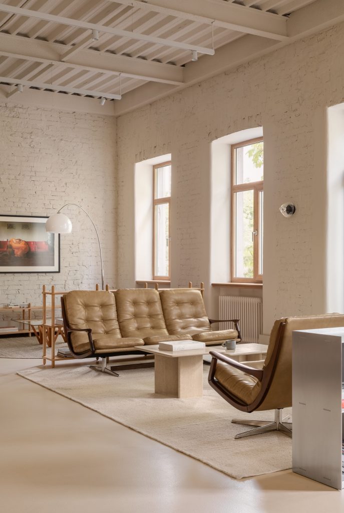 Tan leather sofa and armchair on a pale rug beneath a timber joist ceiling, arched windows in whitewashed brick