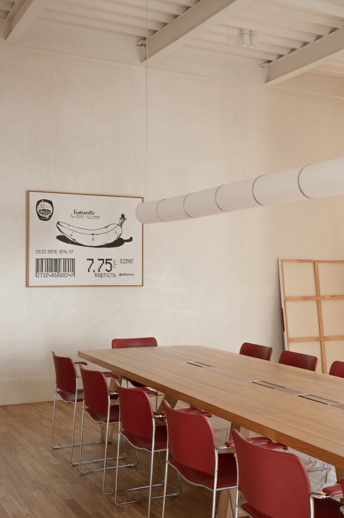 Long oak communal table with red stacking chairs beneath a segmented tube pendant, a banana-receipt artwork on the wall