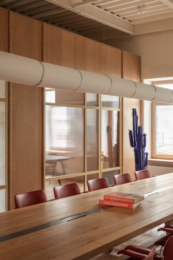 Communal table with red chairs beside fluted-glass and oak partitions, a blue cactus sculpture at the window