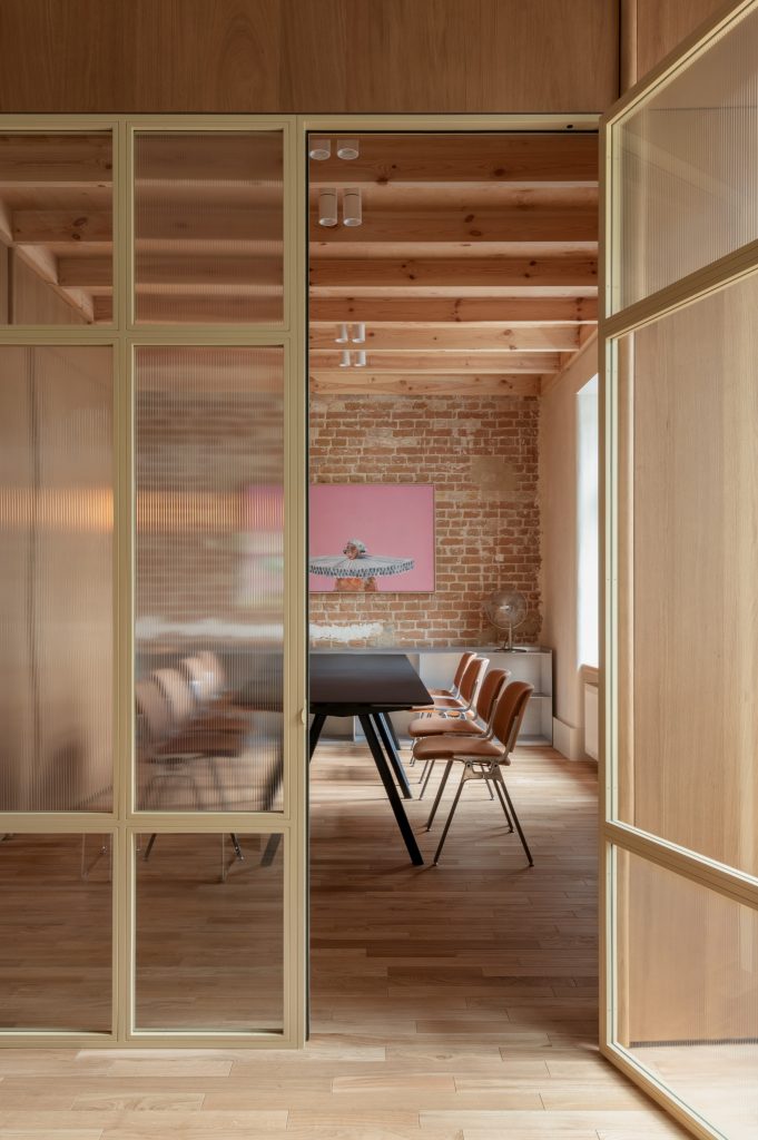 Meeting room seen through brass-framed fluted glass: exposed brick, timber beams, black table, tan chairs and a pink artwork