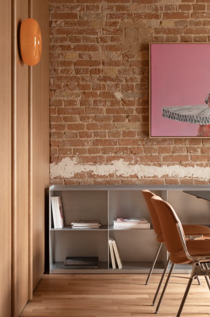 Exposed brick wall with a pink painting, an orange glass sconce and a grey credenza of books below
