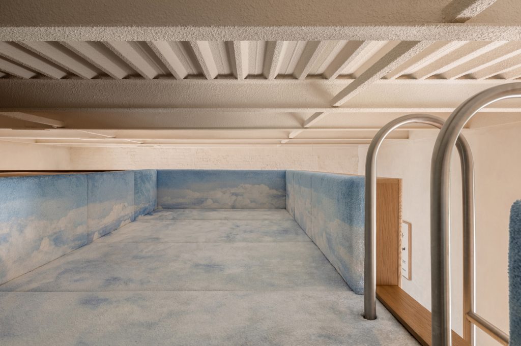 Mezzanine lounge upholstered in sky-printed mats, a steel pool ladder rising to it beneath a white joist ceiling