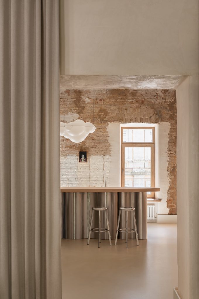View past a curtain to the kitchen: stainless bar counter, cloud-shaped pendant and raw brick behind