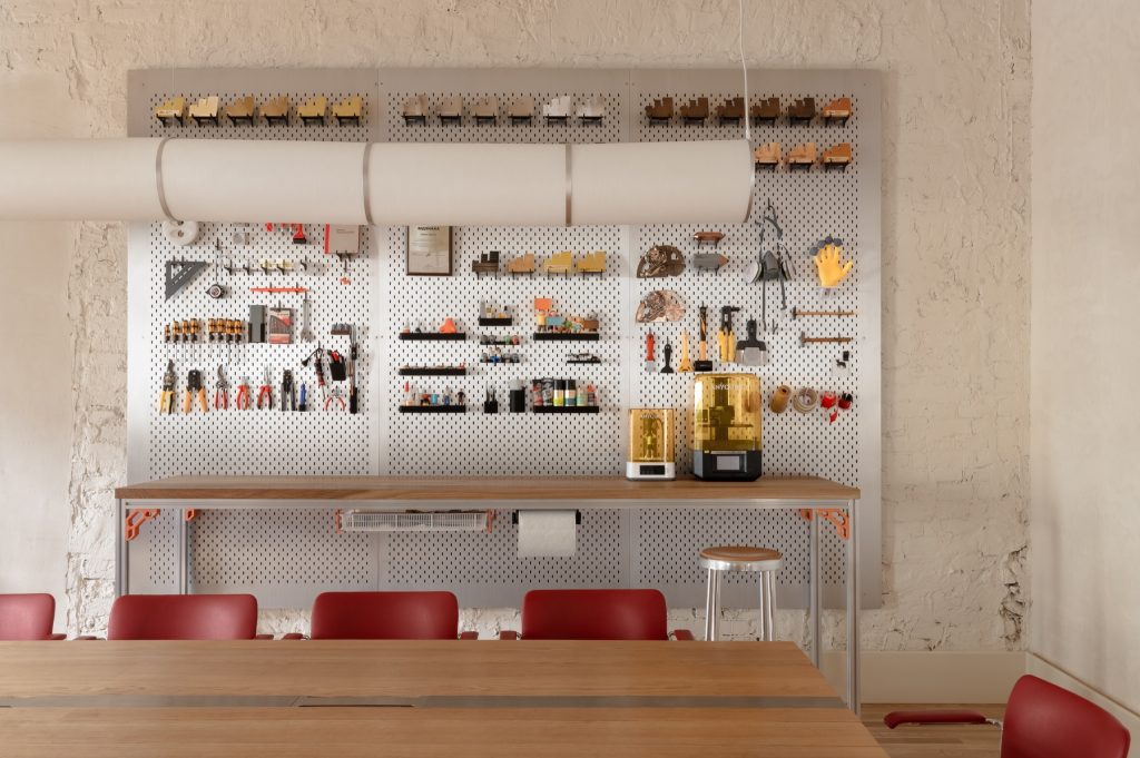 Brushed aluminium pegboard wall hung with tools, model chairs and awards above a long worktable with red chairs