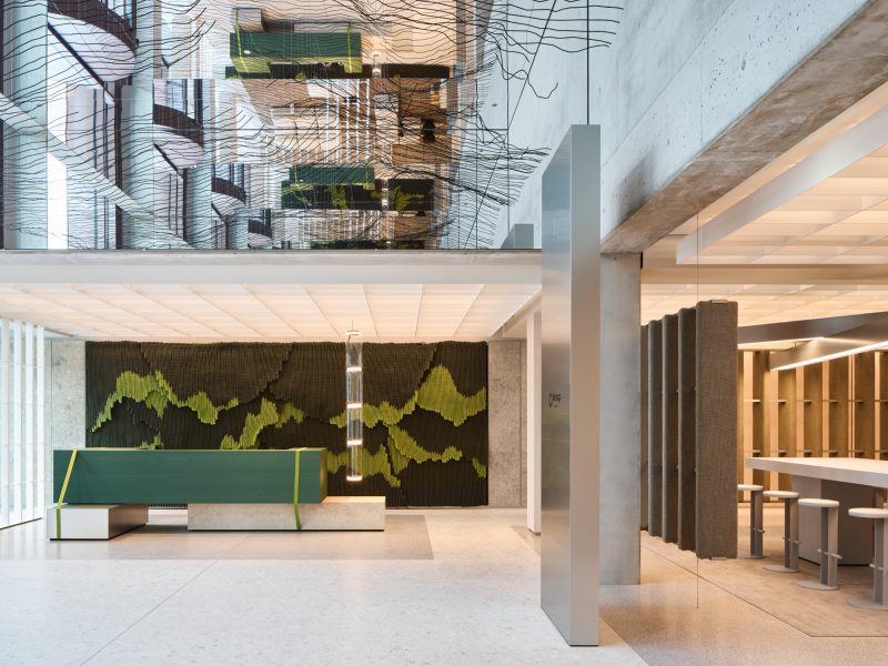 Lobby with a green reception desk strapped like a shipping crate, a woven rope artwork behind and the wire wave mirrored above
