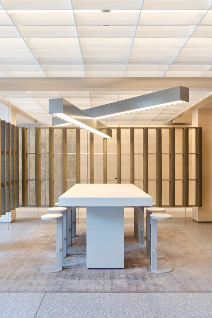 High table and stools beneath a crossed aluminium light, fabric baffles and limestone beyond