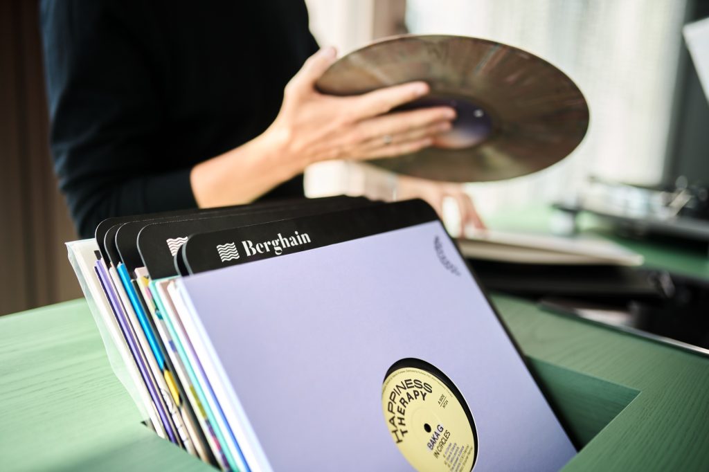 A crate of records topped by a Berghain sleeve, a hand lifting a disc toward the player