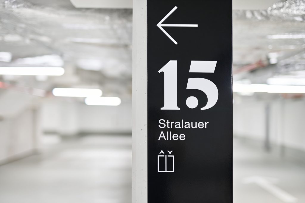 Black wayfinding pylon reading 15 Stralauer Allee with lift pictogram, in the white parking garage