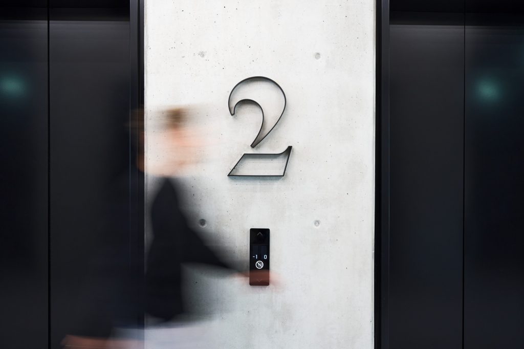 A bent-steel numeral two mounted on board-marked concrete beside the lifts, a person passing in blur
