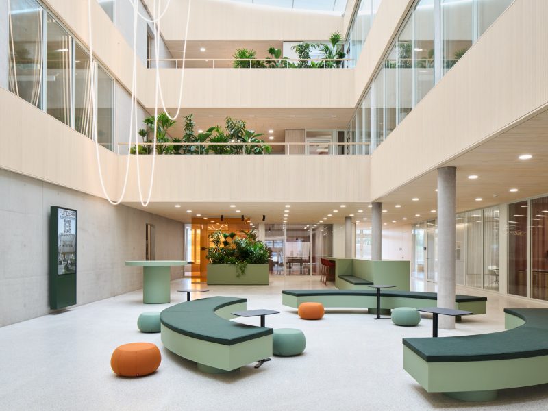 Atrium floor with curved green benches, orange poufs and a tubular light sculpture hanging through three planted storeys