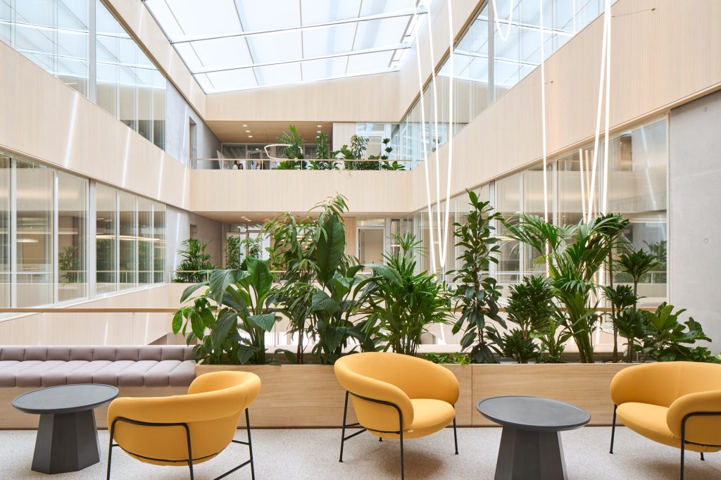 Atrium seen from a balcony: glazed roof above, dense planting on the parapets, yellow armchairs and a pink banquette below