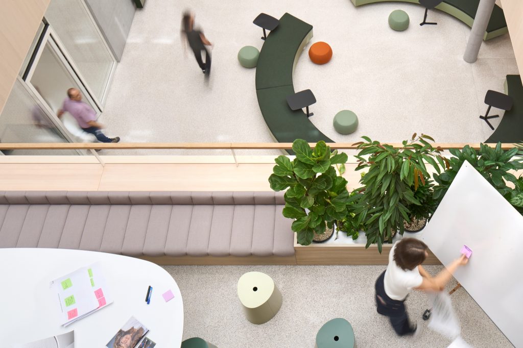 Looking down into the atrium: a green curved bench, poufs, planted parapet and people crossing the terrazzo floor