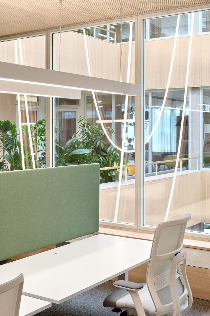 Desk with a green acoustic screen and mesh task chair beside atrium glazing, planting and light tubes beyond