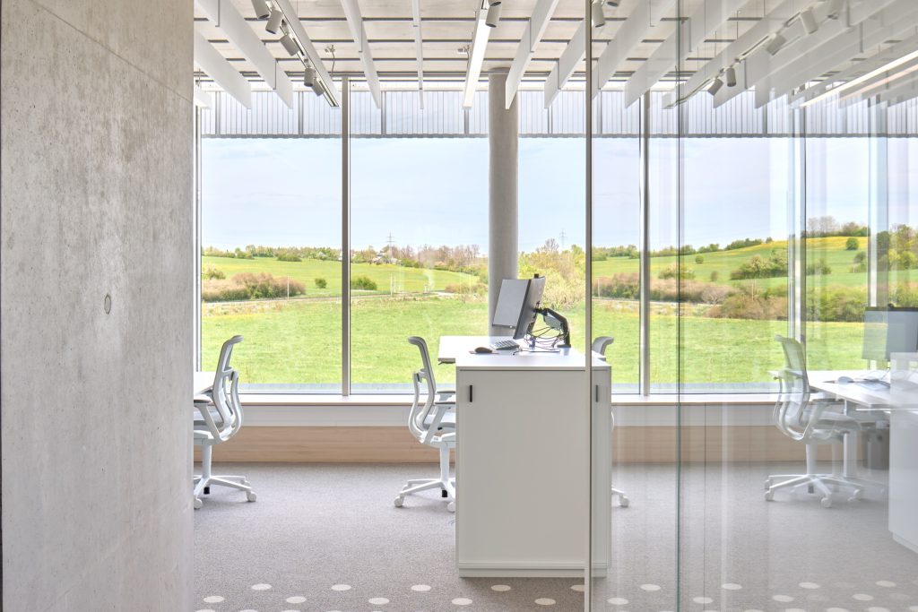 Desks and white task chairs at full-height glazing overlooking meadows and orchards, concrete column alongside