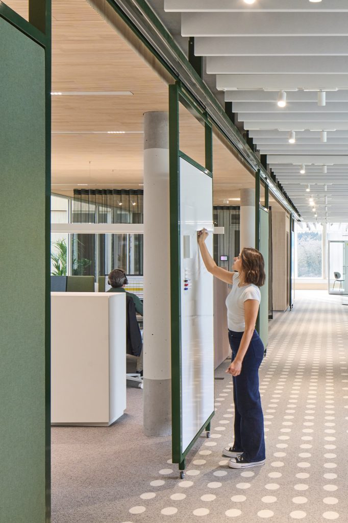 A woman writing on a green-framed mobile whiteboard in a corridor, acoustic baffles overhead and dotted carpet underfoot