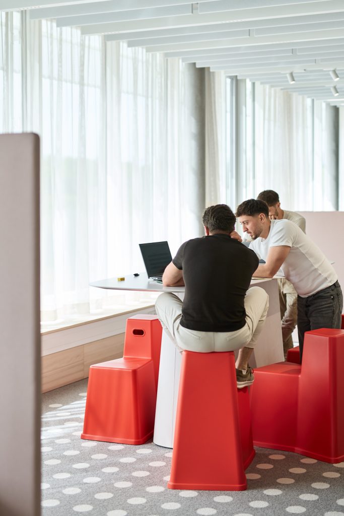 Three people at a high table on red plastic stools in front of sheer curtains and slatted acoustic ceiling