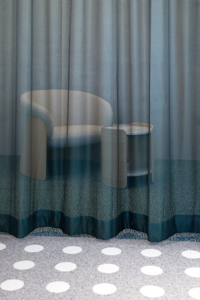 Teal mesh curtain drawn across a meeting area, a chair and table visible through the weave, dotted carpet below
