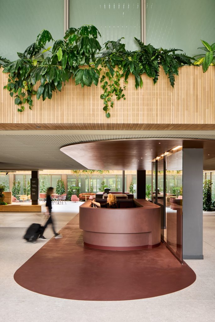 Sculptural oxblood reception counter on a matching floor inset, planting spilling from the slatted timber gallery above