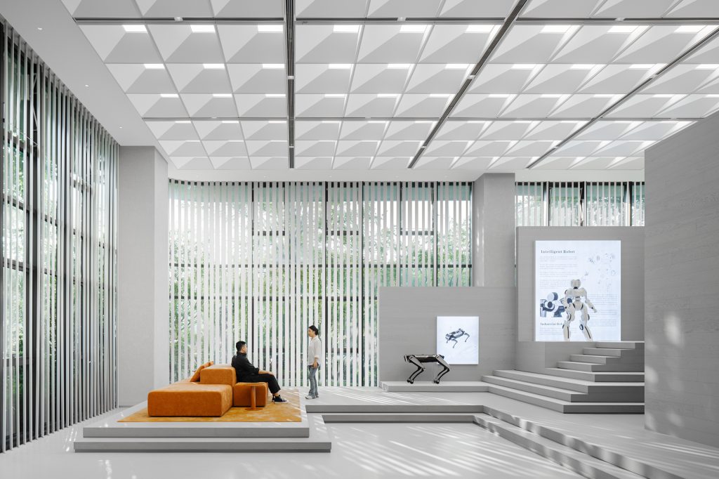 Work lounge and exhibition area beside a louvred glass facade, with an orange modular sofa and robotics displays