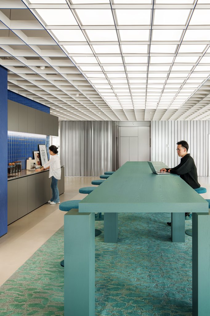 Communal teal standing-height table beneath a luminous grid ceiling, with a blue-tiled tea point