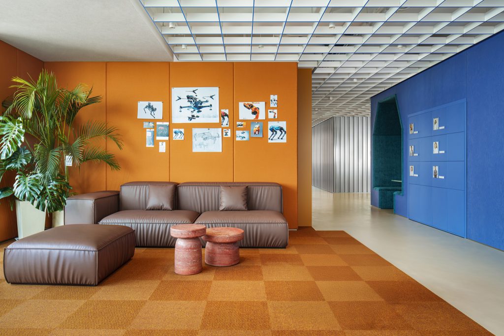 Breakout lounge with a pin-up wall of robotics prints, leather sofas and checkerboard carpet; lockers and a teal booth beyond