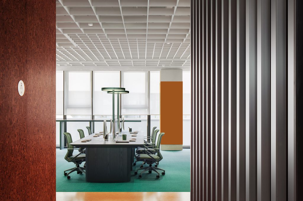 Team workspace with green desks and task chairs on sea-green carpet, seen past a marbled-plywood wall