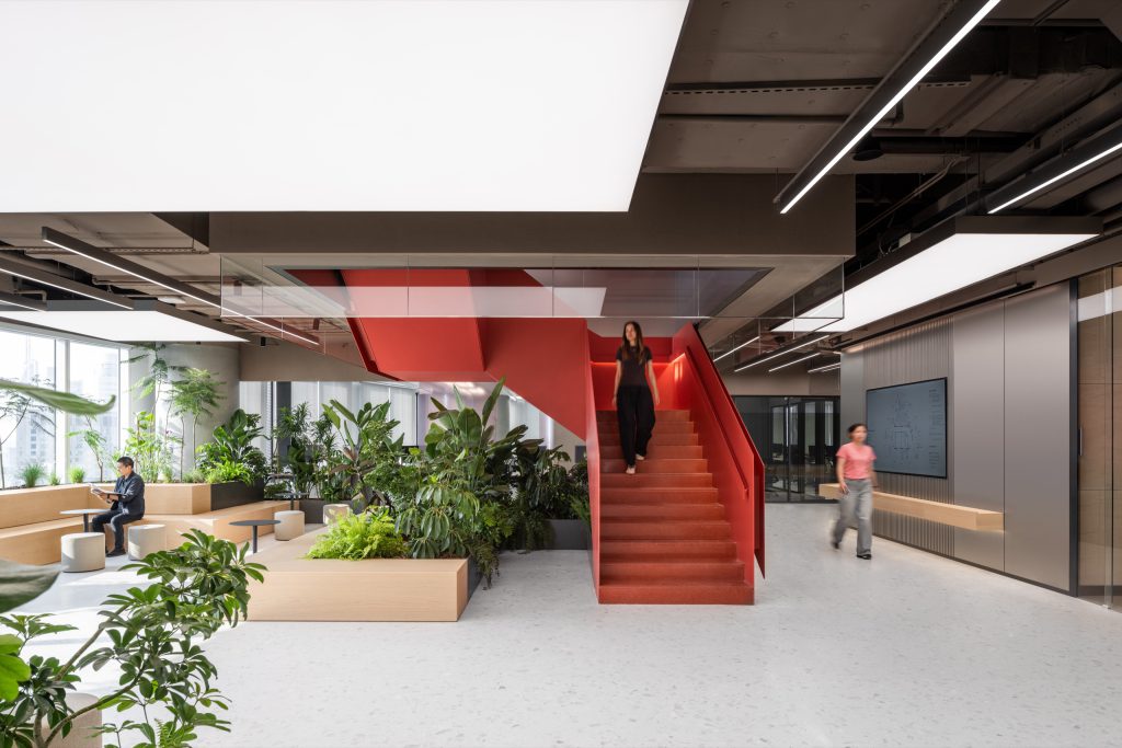 Red sculptural staircase rising through a planted courtyard, beneath luminous ceiling panels