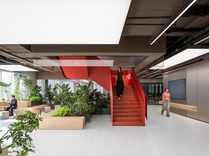 Red sculptural staircase rising through a planted courtyard, beneath luminous ceiling panels