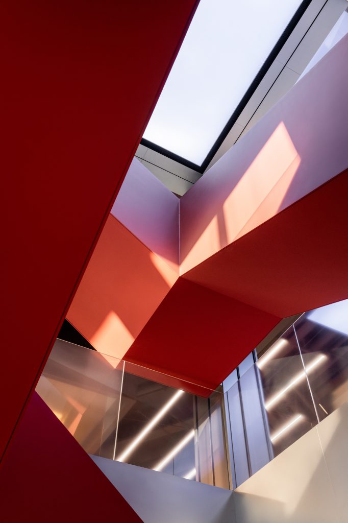 Looking up through the red staircase void toward a luminous ceiling panel