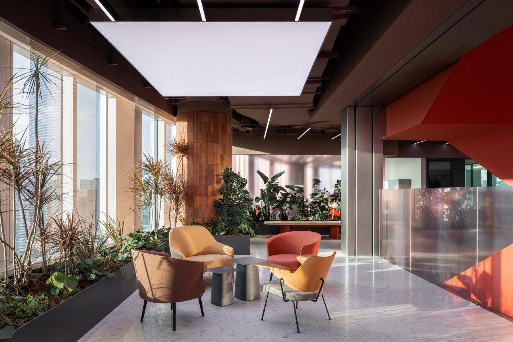 Visitor lounge with coral and ochre armchairs among window planters, beside a brick-clad column