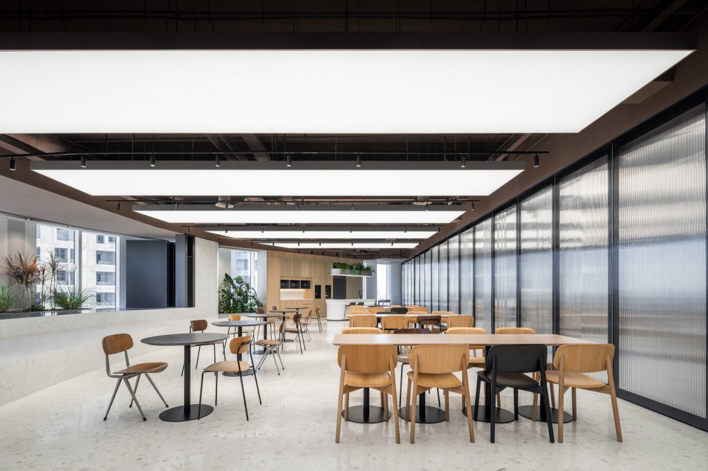 Dining tables beneath luminous ceiling rafts, beside a ribbed-glass wall