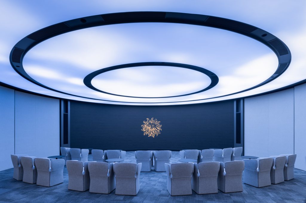 Circular pitch room with a ring-lit sky ceiling, curved screen wall and grey armchairs, the Publicis lion at its centre
