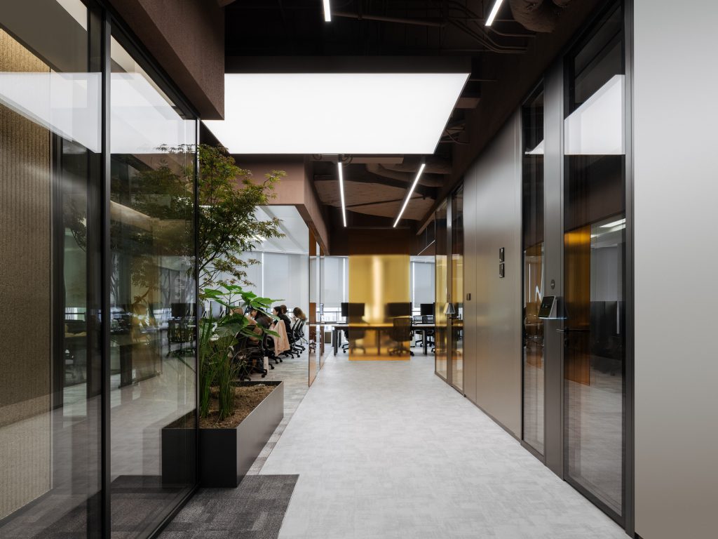 Corridor past glass-fronted meeting rooms toward open desks, an amber glass screen glowing at its end