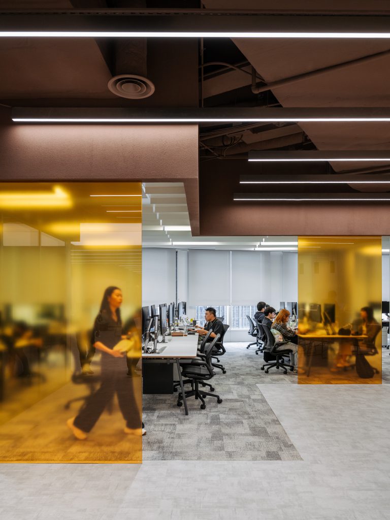 Open workspace with amber glass screens, white desks on red frames and linear lighting overhead