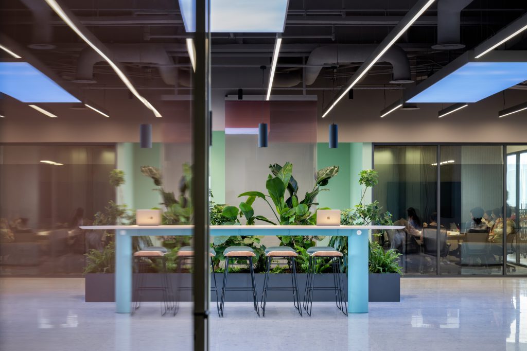 Standing-height table with stools set into a planter between glass meeting rooms, sky panels glowing above