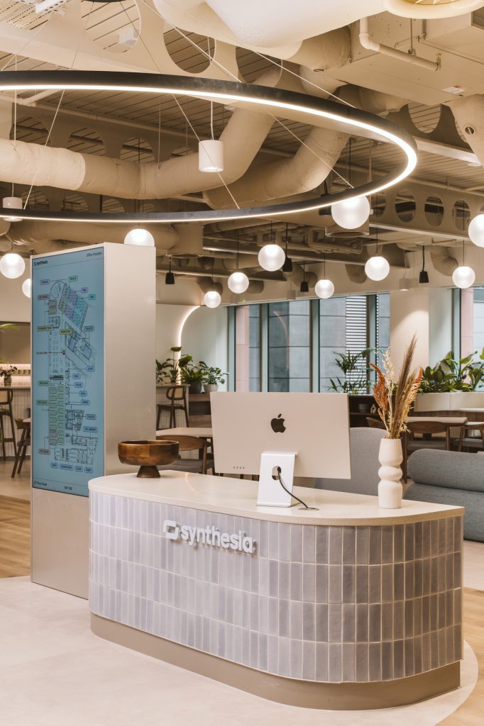 Curved reception desk in pale grey tiles with the Synthesia logo, digital floor directory alongside