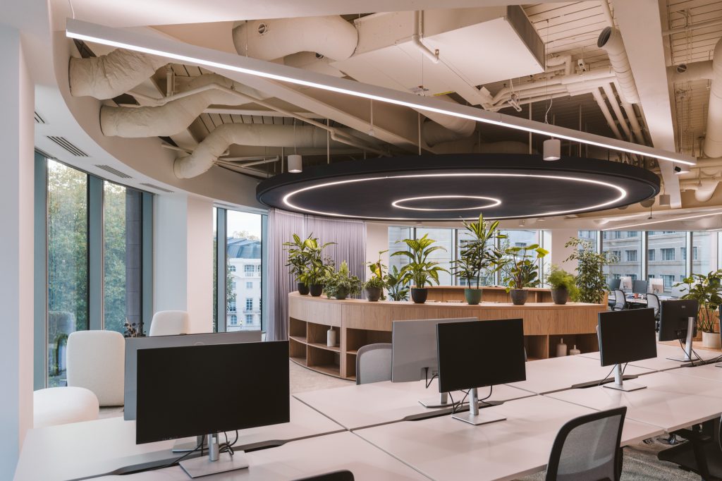 Rows of desks beneath a suspended disc with light rings, a curved planted credenza behind