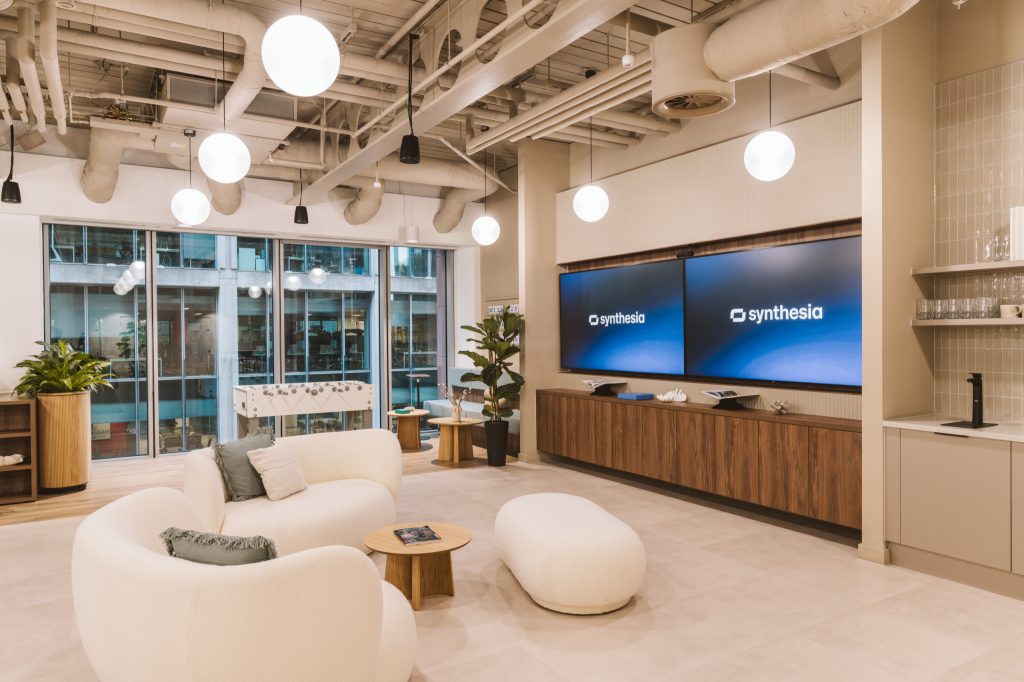 Cream boucle sofas and a pouf facing twin branded screens, a foosball table at the window