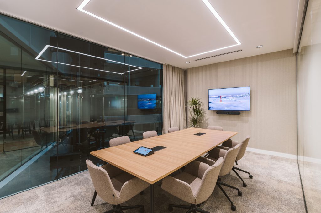 Meeting room with timber table, taupe chairs and a video screen beneath a rectangular light halo