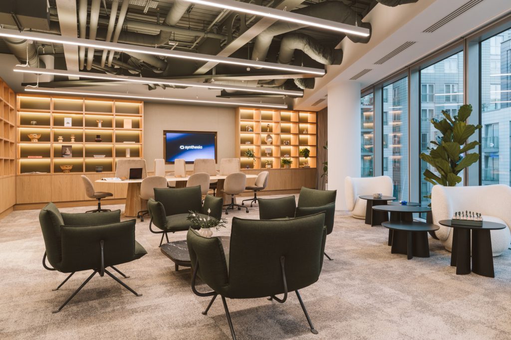 Warm library with backlit oak shelving, deep green lounge chairs and a communal work table